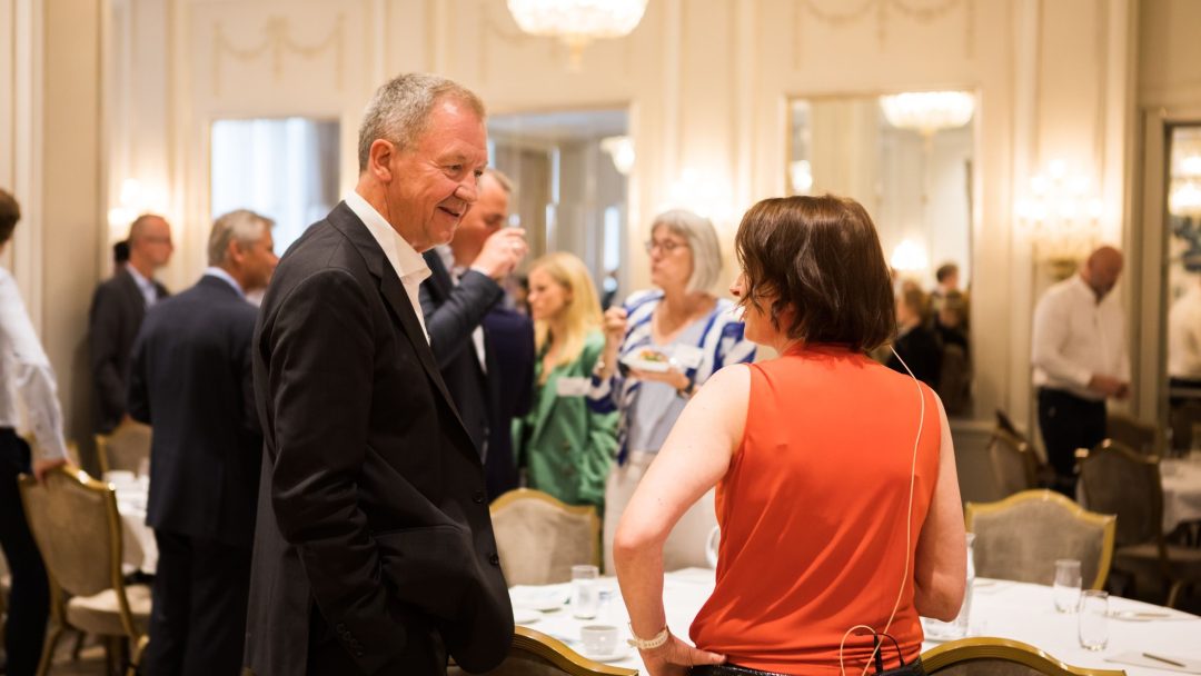 Folk blander seg på et formelt arrangement i et elegant rom med lysekroner. En mann i dress og en kvinne i en rød topp snakker i forgrunnen.