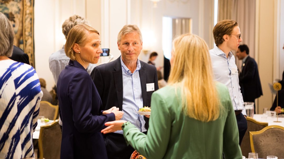 Folk gikk i samtale på et formelt innendørsarrangement, med flere personer som sto rundt bord dekket med glass og tallerkener.