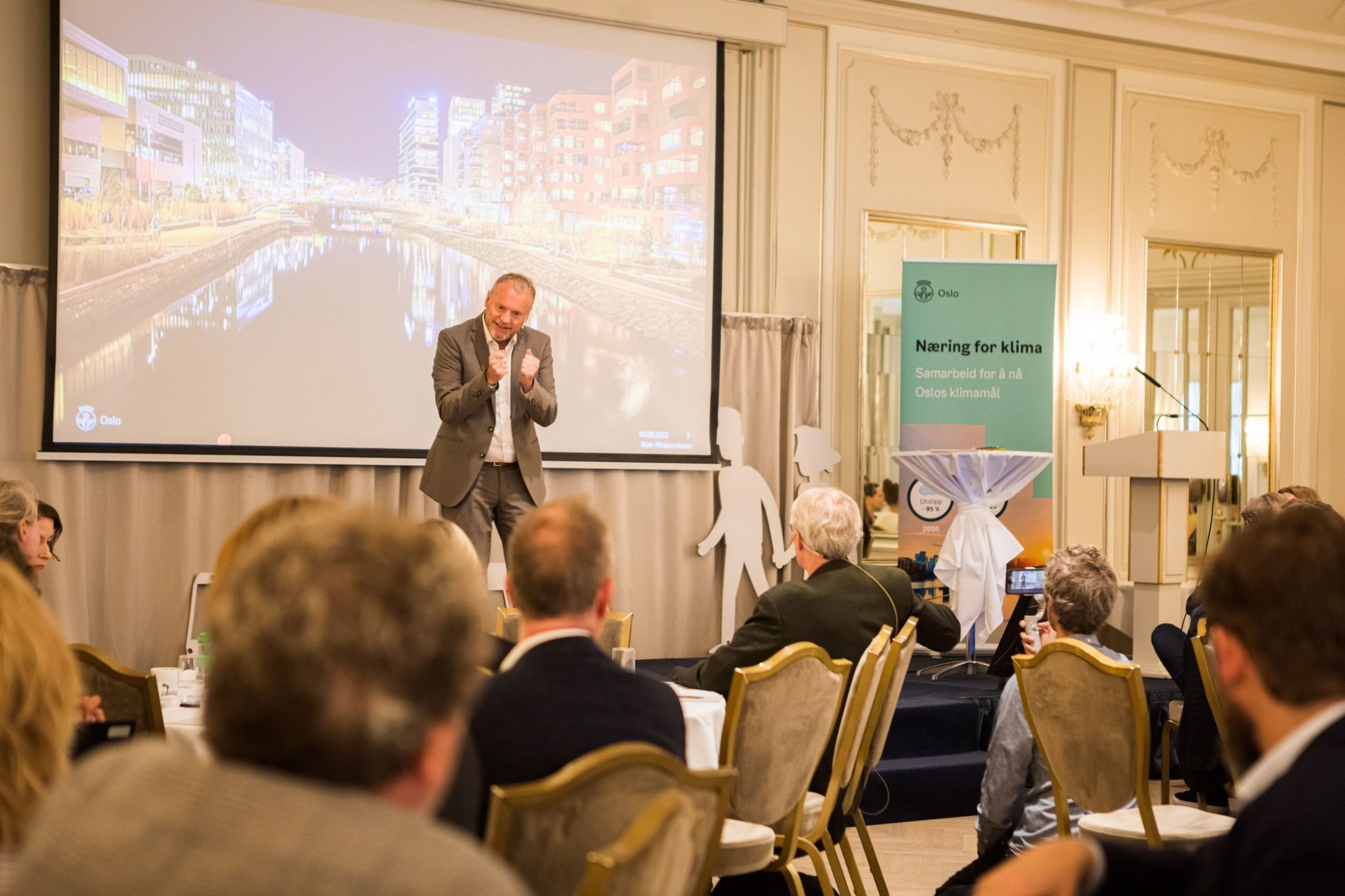 En høyttaler i dress gestikulerer på scenen foran et publikum. En skjerm viser et bybilde, og et banner om klimasamarbeid er synlig.