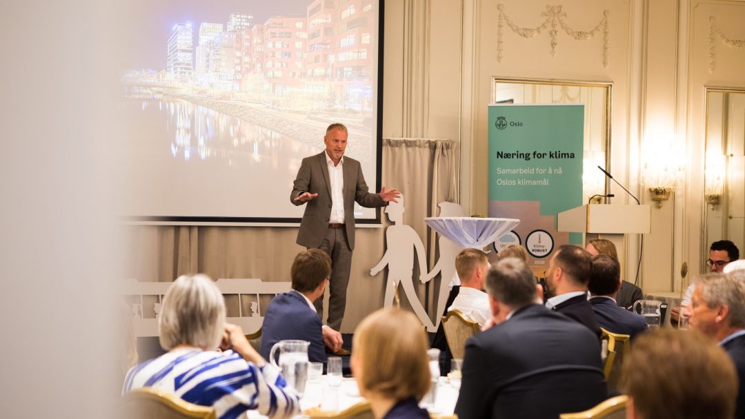 En mann i dress snakker til et publikum på et forretningsarrangement. En projektor viser et bybilde. Et banner med tekst om klimapartnerskap er synlig. Deltakerne sitter vendt mot foredragsholderen.