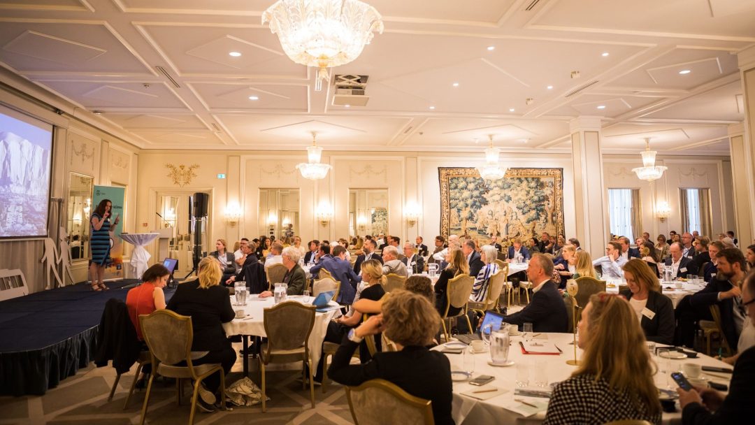 En foredragsholder presenterer på en konferanse, med deltakere ved runde bord i et elegant, lysekroneopplyst rom.