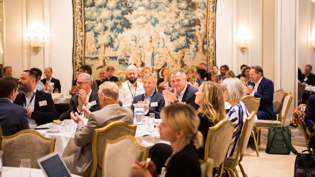 En gruppe mennesker satt ved bordene i et konferanserom, noen klappet. Et stort dekorativt billedvev henger på veggen i bakgrunnen.