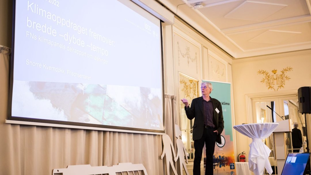 En mann holder en presentasjon i et dekorert rom, og peker på en stor skjerm som viser tekst.