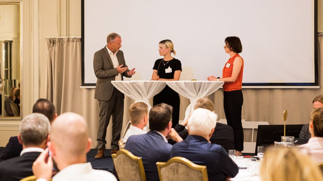 Tre personer på scenen deltar i en paneldiskusjon foran et publikum, med en mann som snakker og to kvinner som lytter.