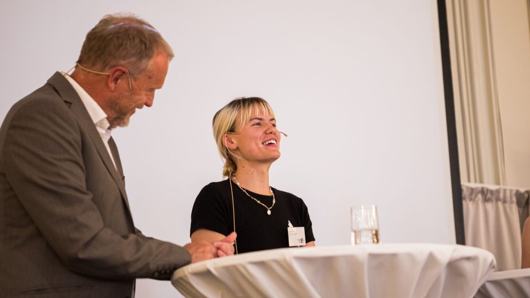 To personer snakker ved et rundt bord med mikrofoner festet, den ene i dress og den andre i en svart skjorte, mot et vanlig bakteppe.