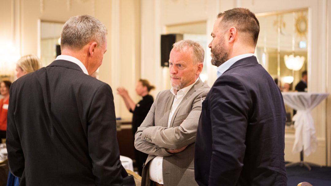 Tre menn i dress gikk i samtale på et innendørsarrangement, med andre deltakere synlige i bakgrunnen.