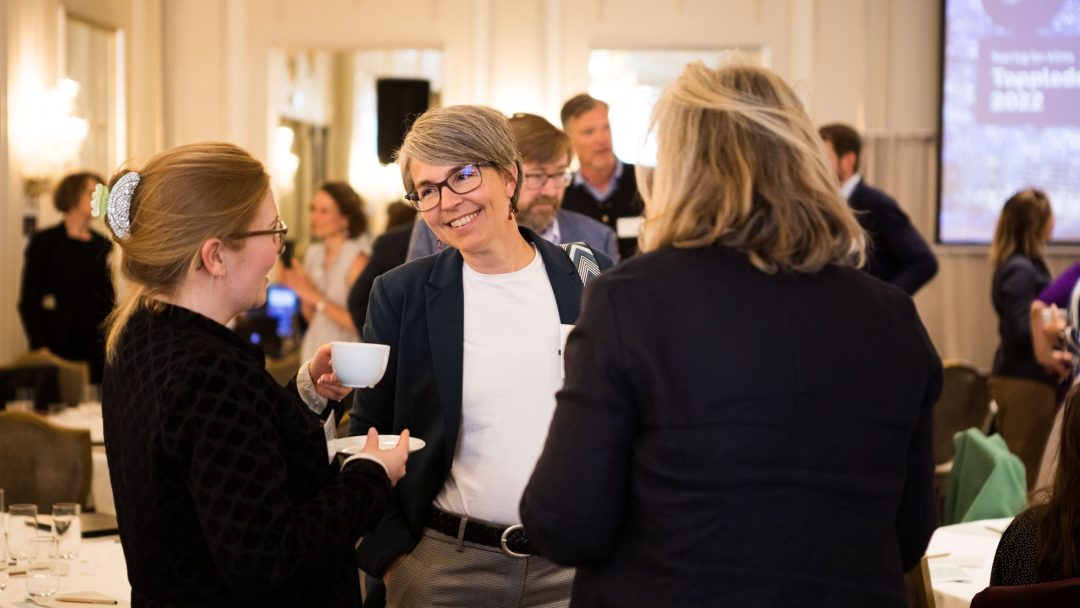 Folk som samhandler på et konferansested, med en person som holder en kopp. Bord, stoler og et projektorlerret er synlig i bakgrunnen.