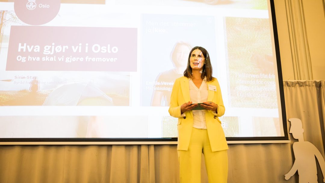 En person i gul dress står på scenen foran en projisert presentasjon og snakker til et publikum.