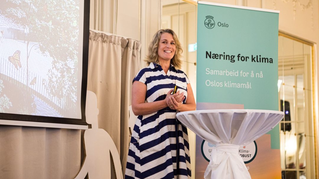 En kvinne i stripet kjole snakker på et podium ved siden av et banner og en skjerm. Innstillingen foreslår en konferanse eller presentasjon.