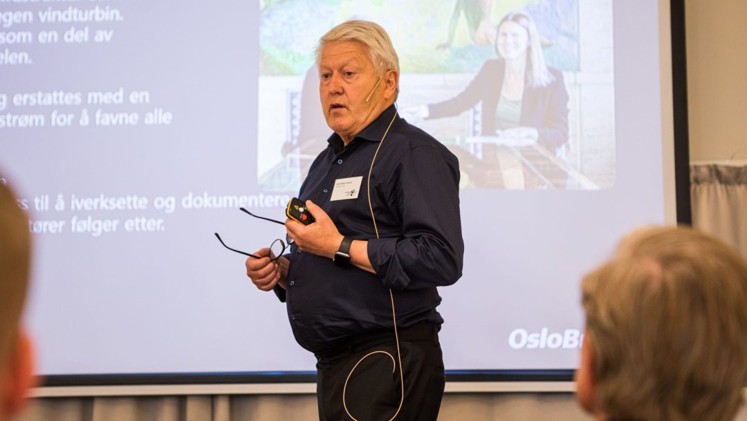 En eldre mann i svart skjorte holder briller og en enhet og snakker foran en presentasjonsskjerm. Publikumsmedlemmer er litt synlige i forgrunnen.