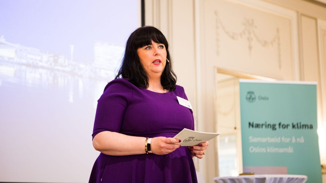 En kvinne i lilla kjole snakker ved et podium innendørs. Et banner knyttet til Oslos klimamål er synlig i bakgrunnen.