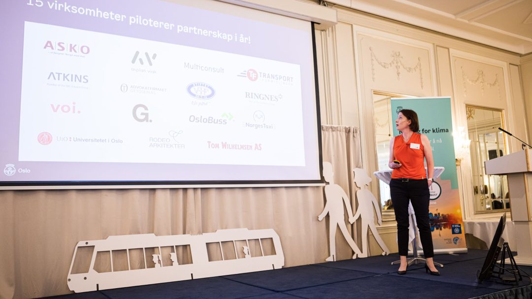 En person står nær en skjerm som viser logoer fra forskjellige selskaper. Innstillingen ser ut til å være en presentasjon i et konferanserom.