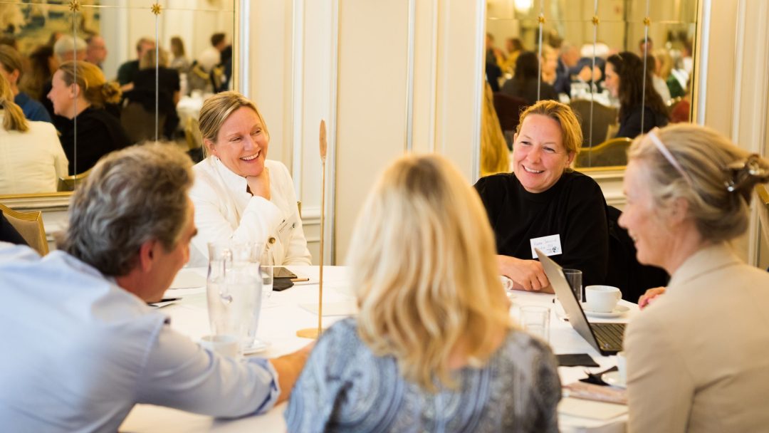 Fem personer sitter rundt et rundt bord i et sterkt opplyst rom, engasjert i samtale, med papirer og en bærbar datamaskin på bordet.