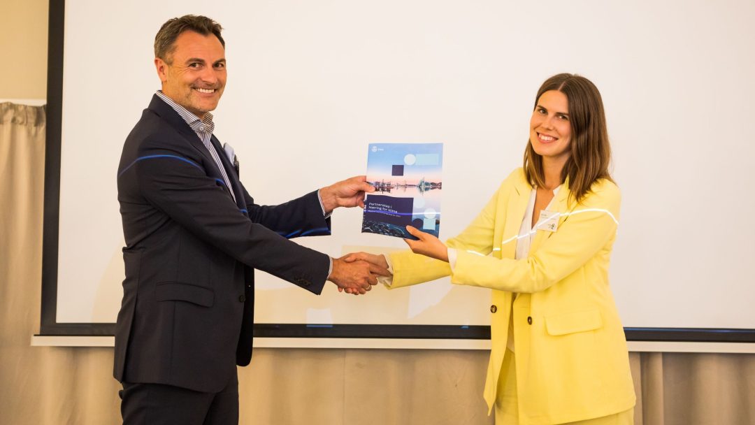 To personer i forretningsantrekk håndhilser og holder et dokument foran en projeksjonsskjerm.