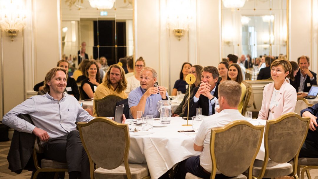 Folk satt rundt et rundt bord og lyttet oppmerksomt i et godt opplyst rom med speil og lysekroner.