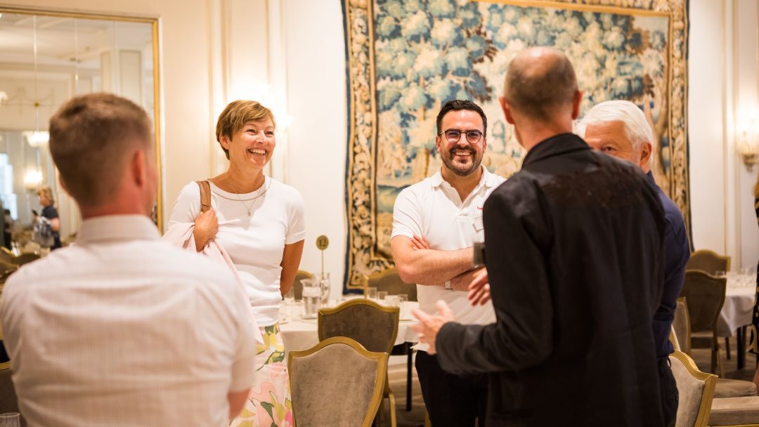 Gruppe på fem personer som står i et rom med elegant innredning, snakker og smiler. Et billedvev henger på veggen i bakgrunnen.