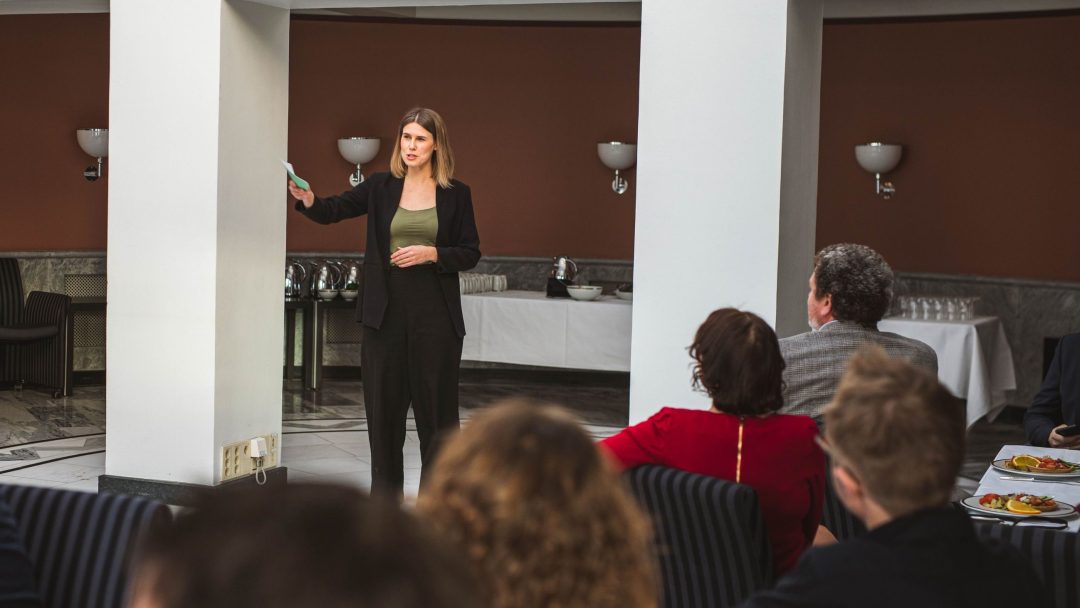 En kvinne i svart dress snakker på et innendørs arrangement. Hun står i nærheten av en gruppe sittende mennesker, med bord og buffetartikler synlig i bakgrunnen.