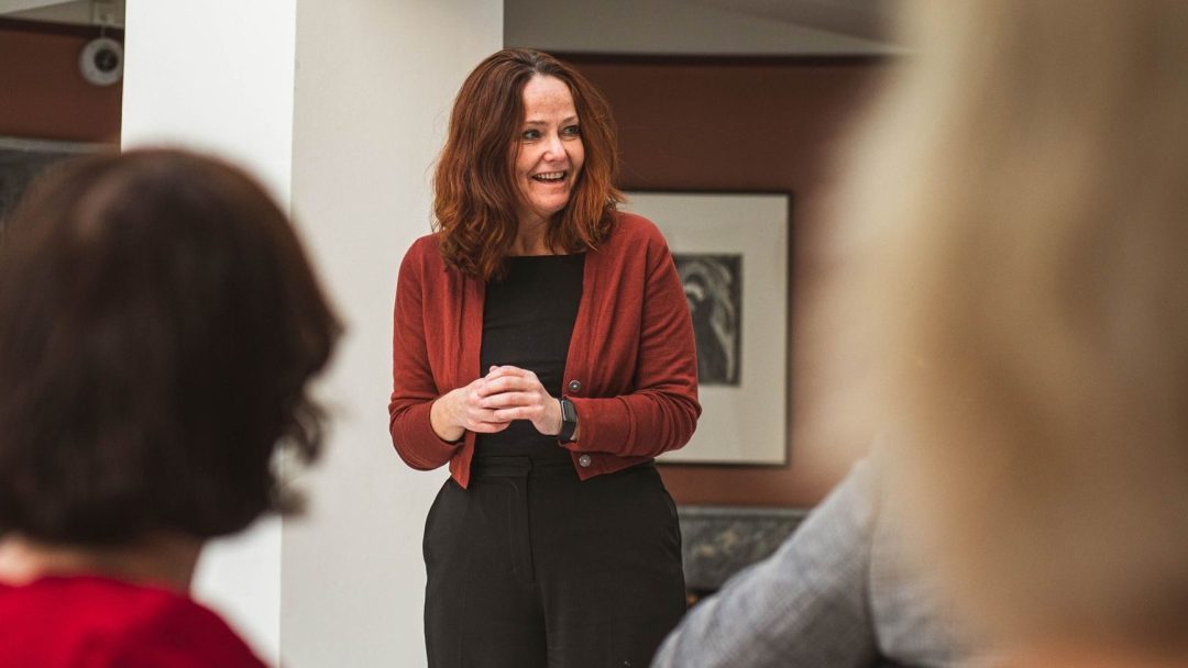 En kvinne med skulderlangt brunt hår, iført en rød cardigan, står og snakker til en liten gruppe mennesker inne i et rom.