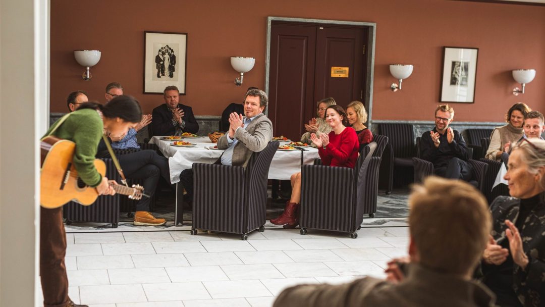 En person som spiller gitar mottar applaus fra et sittende publikum i et rom med innrammede bilder på veggene.