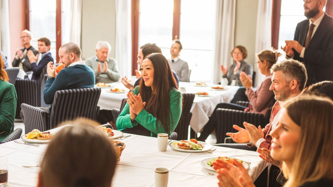 En gruppe mennesker sitter ved bordene i et sterkt opplyst rom og klapper. Bord har tallerkener med mat og drikke.