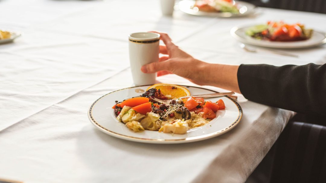 En person strekker seg etter en kopp kaffe ved siden av en tallerken med salat, grillede grønnsaker og ost på en hvit duk.