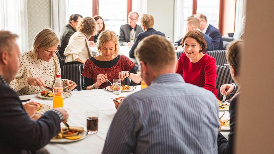 En gruppe mennesker som sitter ved et bord og spiser et måltid sammen, med tallerkener og drinker på bordet i et lyst rom.