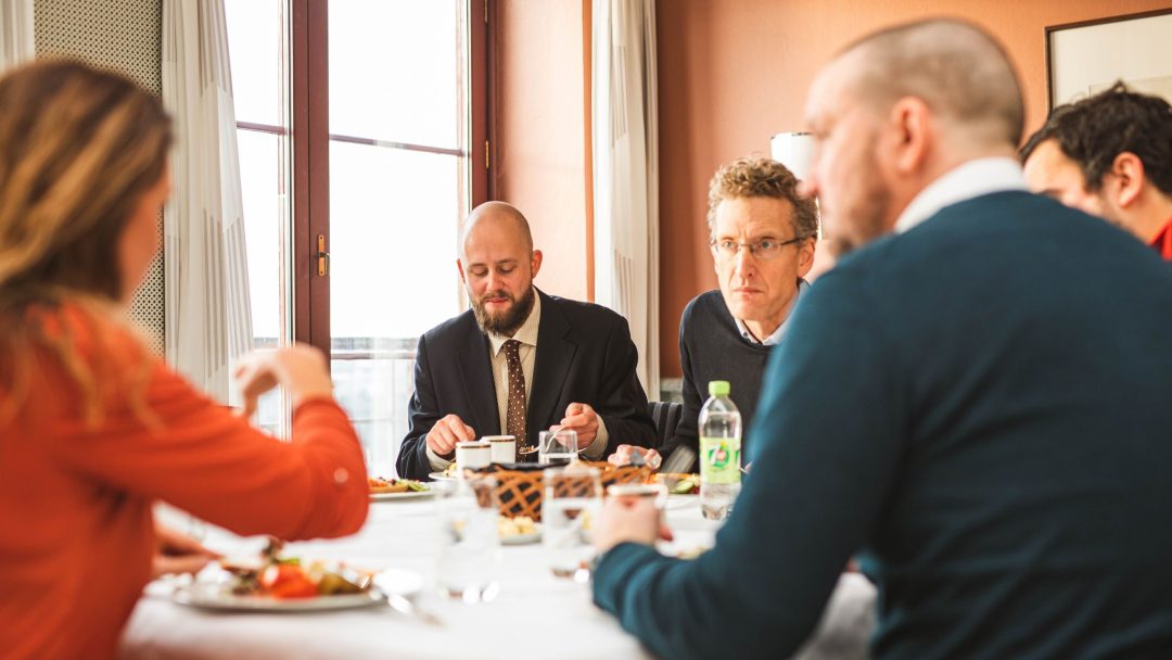 Fem personer sitter rundt et bord og spiser og snakker i et rom med varm belysning. Et vindu er i bakgrunnen. Én person har en notisblokk. En flaske drikke står på bordet.