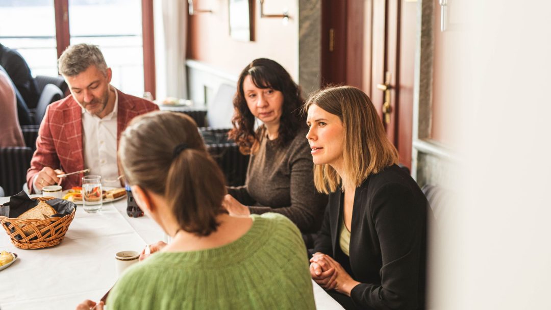 Fire personer sitter ved et bord på en restaurant, engasjert i samtale. En kurv med brød og drikke står på bordet.