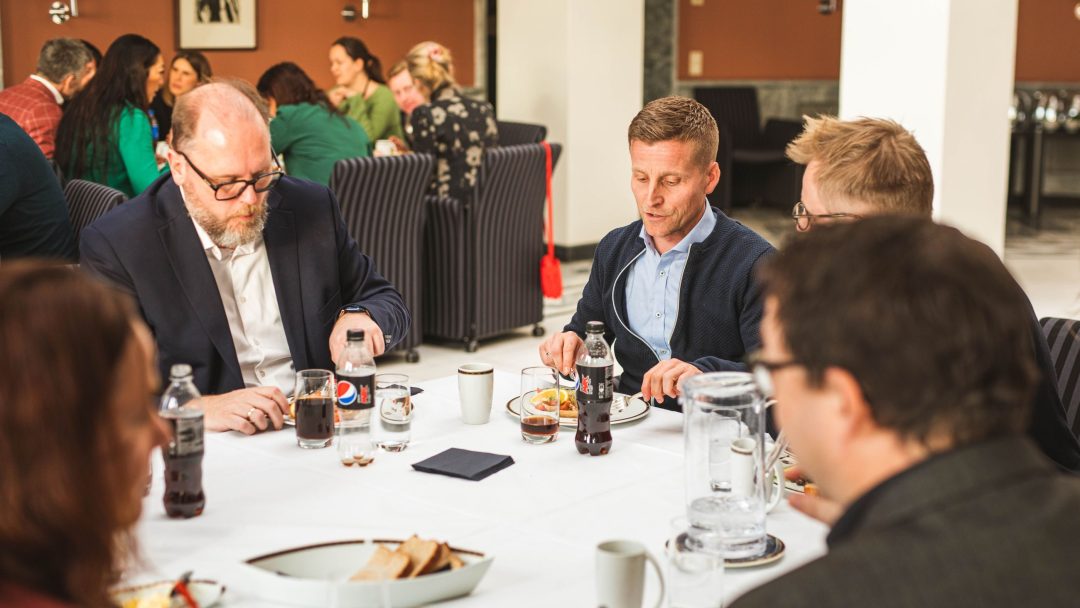 En gruppe mennesker sitter ved et bord med drikke og mat, engasjert i samtale.