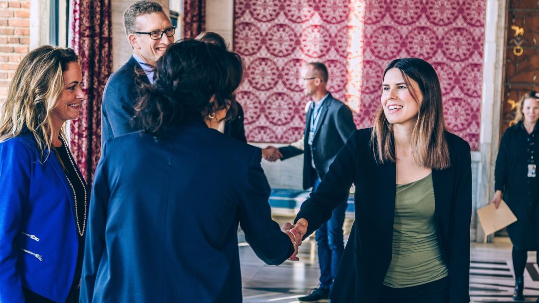 Folk i forretningsantrekk smiler og håndhilser i et varmt innredet rom, med røde mønstrede gardiner i bakgrunnen.