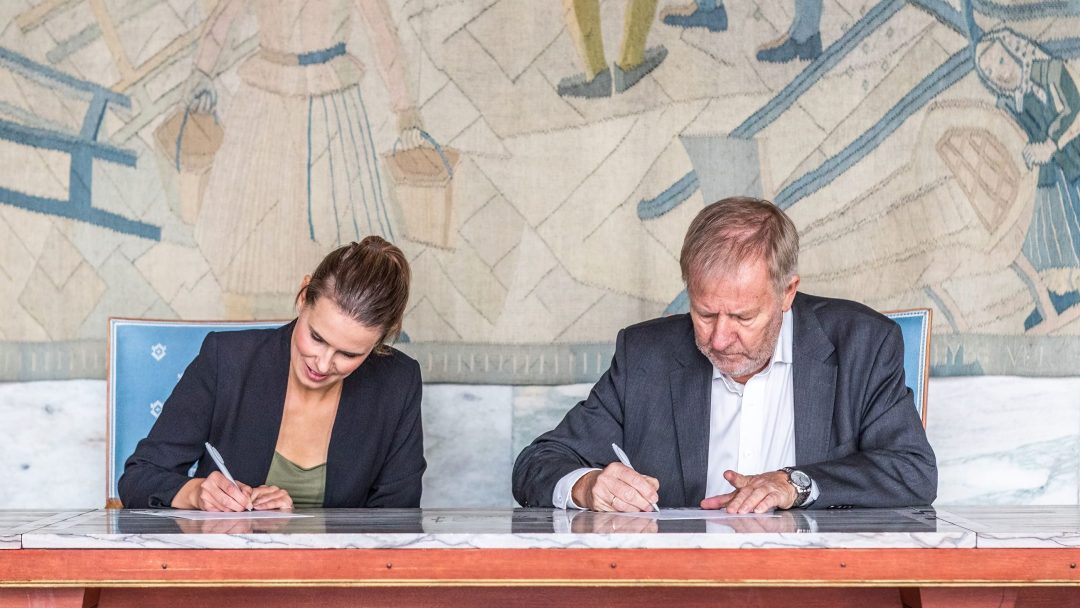 To personer sitter ved et bord og signerer dokumenter. Et billedvev med abstrakte figurer henger i bakgrunnen.