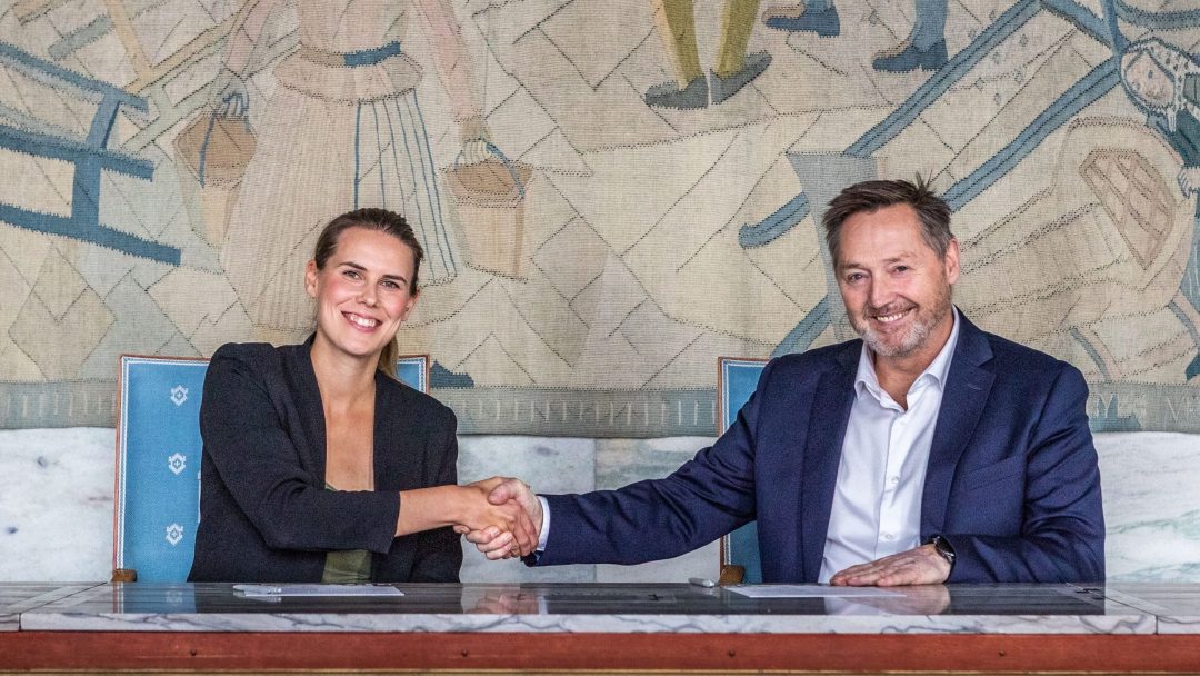 To personer sitter ved et bord og håndhilser og smiler til kameraet. Et billedvev med abstrakte figurer henger på veggen bak.