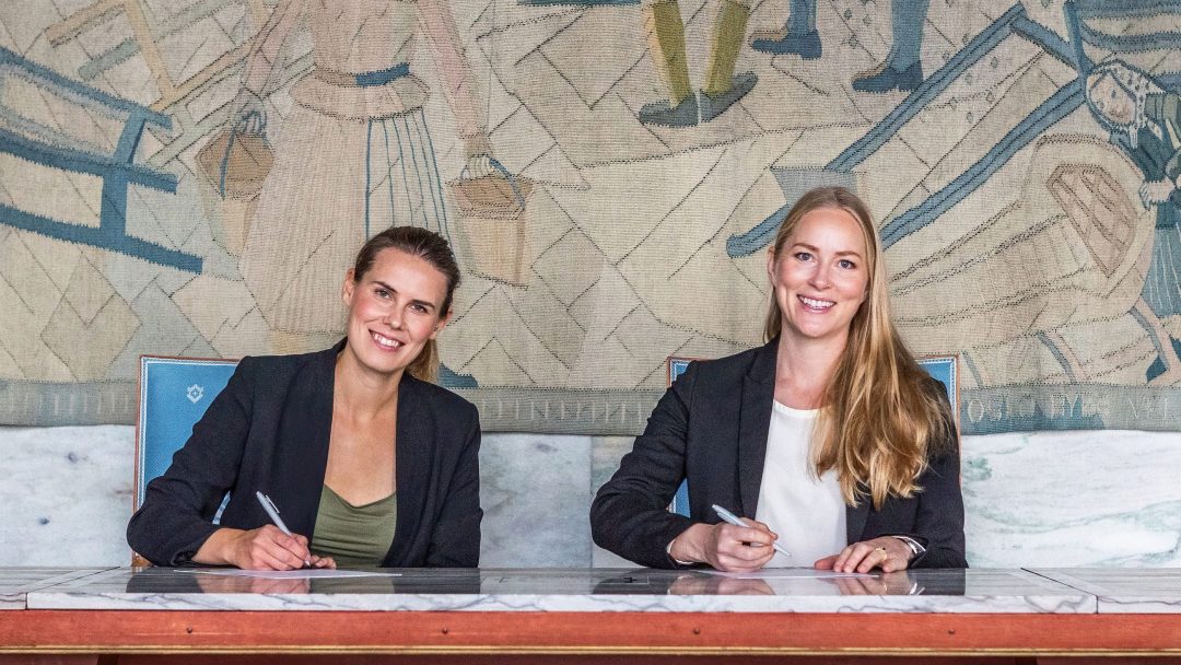 To personer i formell antrekk sitter ved et bord, med hver sin penn over papirer, foran et veggmaleri. De smiler begge til kameraet.