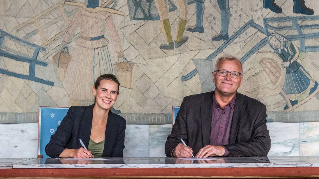 To personer som sitter ved et bord, begge smilende og holder penner, signerer et dokument. Et billedvev med figurer i bakgrunnen pryder veggen.