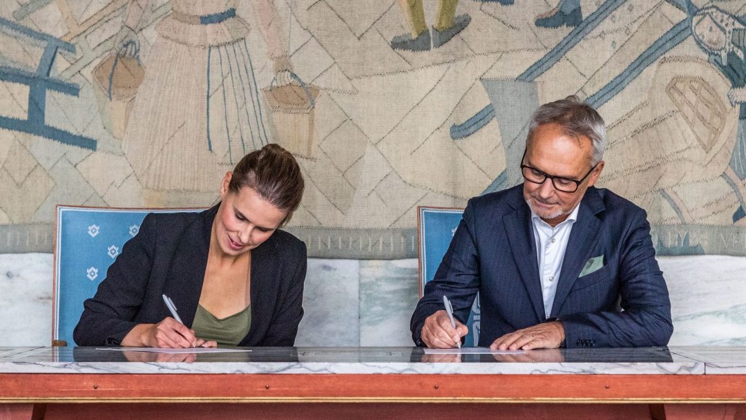 To personer sitter ved et bord, begge signerer dokumenter med penner. Et dekorativt veggmaleri danner bakteppet.