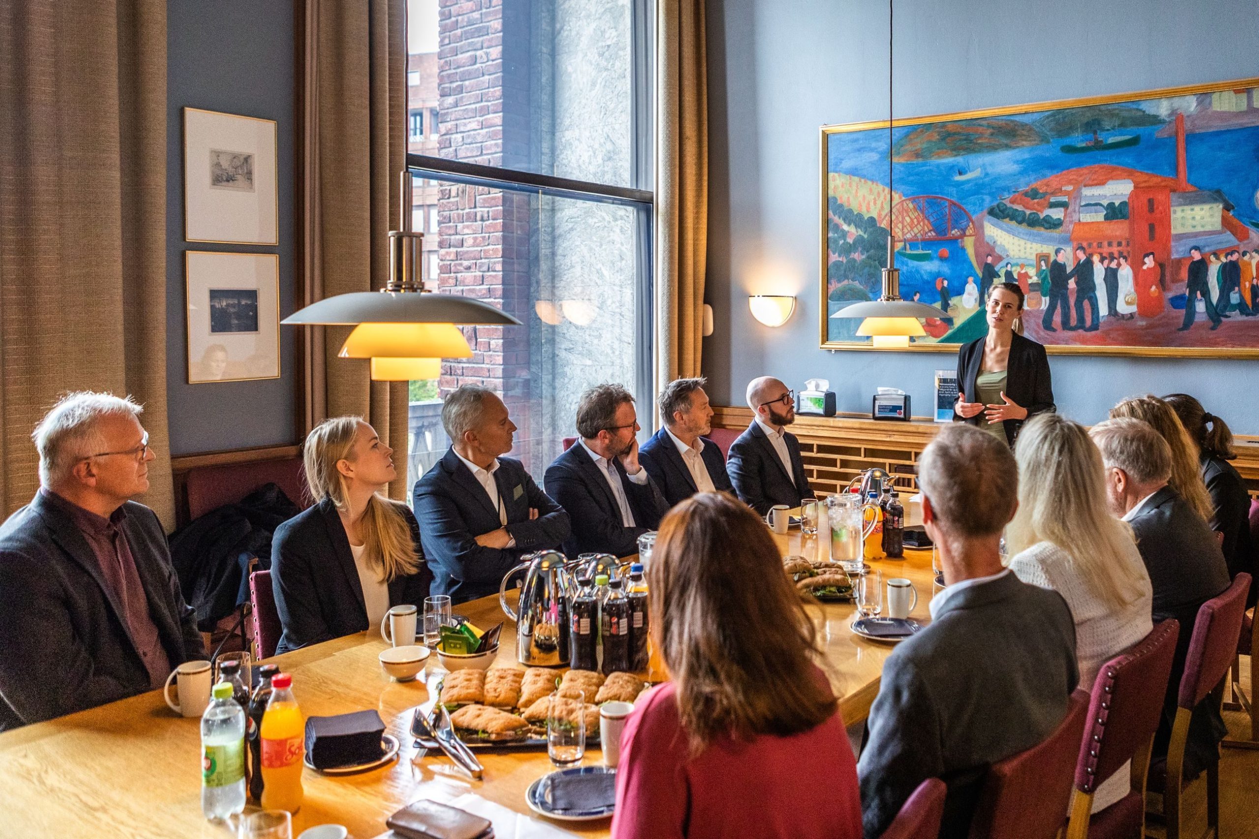 En gruppe mennesker sitter rundt et langbord med drinker og snacks, og hører på en stående høyttaler i et rom med kunstverk på veggen og store vinduer.