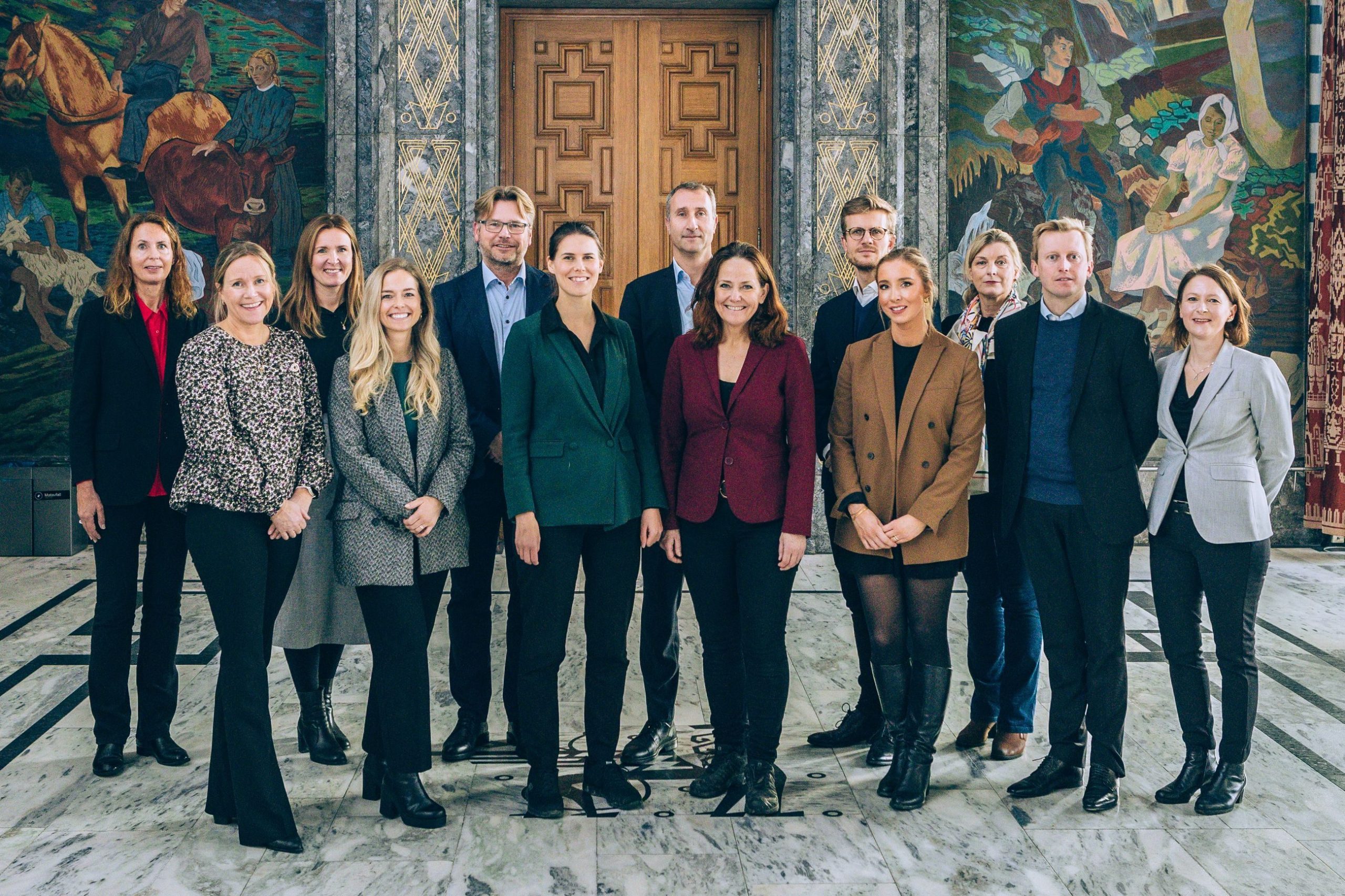 En gruppe på tretten personer i formell antrekk står i et rom med utsmykkede vegger og en mønstret tredør.