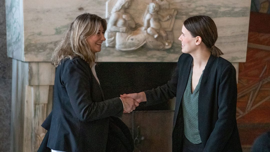 To personer i forretningsantrekk håndhilser i et rom med et marmorrelieff i bakgrunnen.
