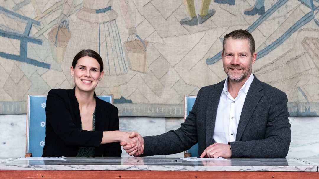 To personer i formell antrekk sitter ved et bord og håndhilser, med et veggmaleri i bakgrunnen.