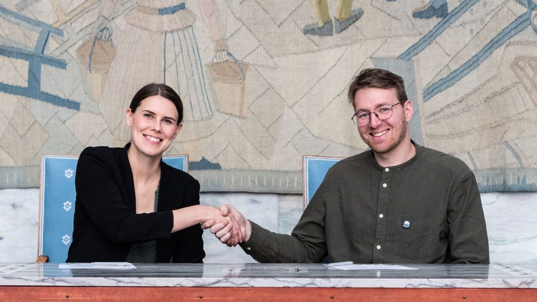 To personer som sitter og håndhilser over et bord, smiler, med et billedvev i bakgrunnen.