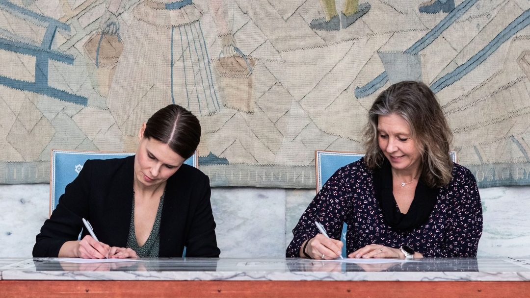 To kvinner sitter ved et bord og signerer dokumenter. Et billedvev med abstrakte figurer henger i bakgrunnen.