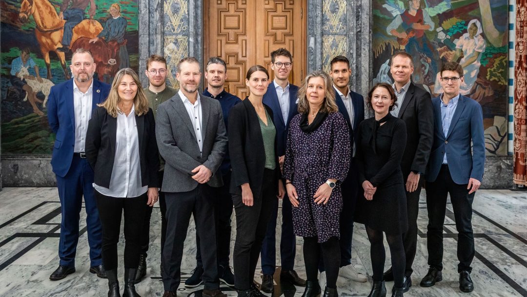 En gruppe på elleve personer står innendørs på et marmorgulv, kledd i forretningsantrekk. Et detaljert veggmaleri er på veggen bak dem.