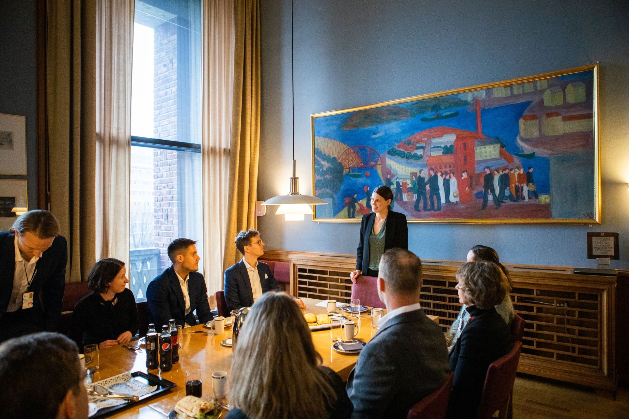 En gruppe mennesker satt rundt et konferansebord og lyttet til en person som står og snakker, med et stort fargerikt maleri på veggen i et rom med høye vinduer.