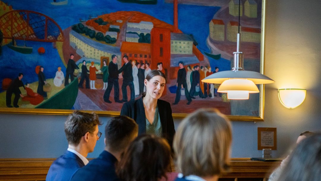 En person står og snakker til en gruppe som sitter rundt et bord i et rom med et stort fargerikt maleri på veggen.