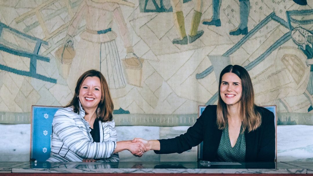 To kvinner som sitter ved et skrivebord, smiler og håndhilser foran et billedvev som bakteppe.