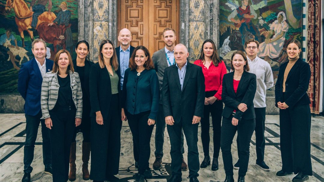 En gruppe på elleve personer står innendørs på et marmorgulv foran en stor tredør, med fargerike veggmalerier på veggene.