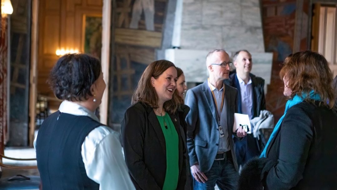 Gruppe mennesker engasjert i samtale i et elegant rom med veggmalerier og klassisk dekor.