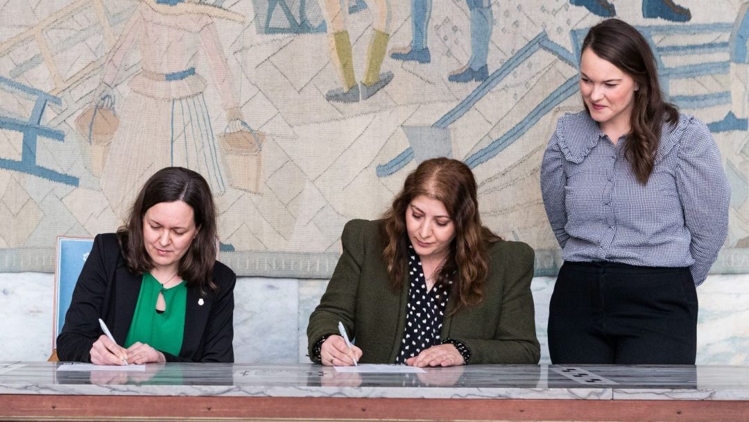 Tre kvinner sitter og står ved et bord og signerer dokumenter, med et stort billedvev i bakgrunnen.