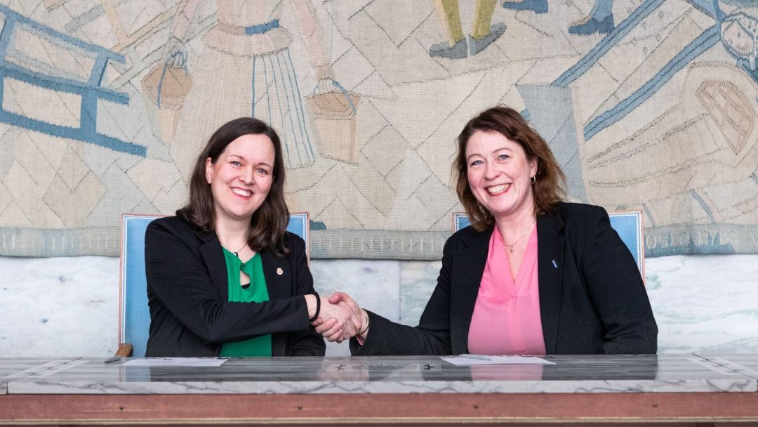 To kvinner som sitter ved et bord håndhilser og smiler. Et billedvev med abstrakte figurer henger i bakgrunnen.