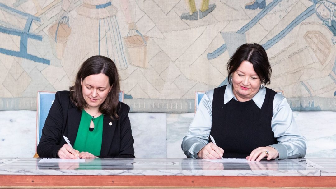 To kvinner sitter ved et bord og signerer dokumenter. Bakgrunnen har et detaljert veggmaleri.